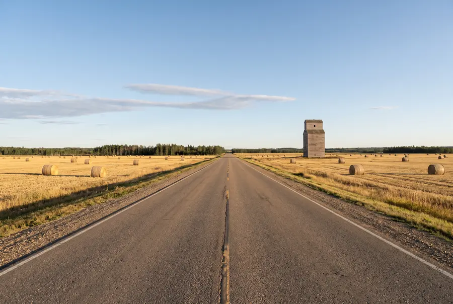 Generated illustration: a straight two-lane highway through harvested fields with hay bales and an old wooden grain elevator; not a specific road.